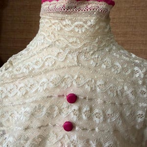 May include: Close-up of an ivory lace garment with a high, ruffled collar. Two magenta buttons fasten the front. The lace features intricate floral patterns, and the collar is trimmed with a magenta fabric.