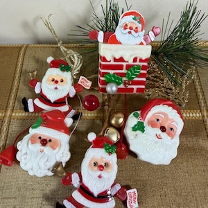 May include: A collection of five vintage-style plastic Santa Claus Christmas ornaments. The set includes figures of Santa emerging from a brick chimney, Santa faces, and full-body Santa figures holding small bells and signs that read Merry Christmas.