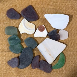 May include: An assortment of sea glass pieces in various shapes and colors, including green, purple, white, and black. A penny is placed on a white piece of sea glass for size comparison. The pieces are arranged on a textured, tan surface.
