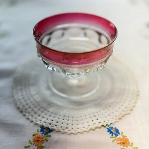 May include: A clear glass dessert dish with a ruby red rim. The dish sits on a white crocheted doily, with a floral embroidered tablecloth in the background. The dish has a decorative, scalloped edge.