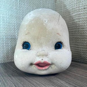 May include: A vintage doll head with a cracked surface. The doll head has large blue eyes, painted eyelashes, and a pink mouth. The head is off-white and sits on a wooden surface.