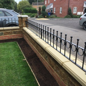 Puede incluir: Una valla de metal negro con elementos decorativos redondeados se asienta sobre un muro de piedra. El muro está hecho de ladrillos de color marrón claro y rojo. La valla está unida al muro con soportes de metal negro.