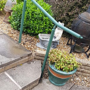 Puede incluir: Una barandilla de metal verde instalada en escalones de hormigón. La barandilla curva está sostenida por dos postes verticales. Una planta en maceta con hojas verdes y rojas está cerca. El fondo incluye vegetación y adornos de jardín.