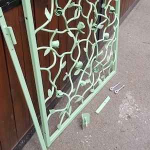 May include: A light green metal gate with a decorative design of roses and vines. The gate is disassembled and laid out on a grey tarmac surface.