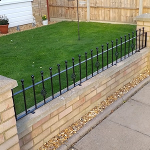 Puede incluir: Una valla de metal negro con remates decorativos se asienta sobre un muro de ladrillo. La valla está unida al muro con soportes metálicos. El muro está hecho de ladrillos de color marrón claro y tiene una tapa de hormigón. El muro está rodeado de un césped verde.