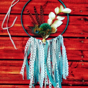 K&ouml;nnte beinhalten: Ein t&uuml;rkisfarbener Traumf&auml;nger mit wei&szlig;er und blauer Spitzenborte, der an einer roten Holzwand h&auml;ngt. Der Traumf&auml;nger ist mit Federn, getrockneten Bl&auml;ttern und einer kleinen gr&uuml;nen Bl&uuml;te verziert.