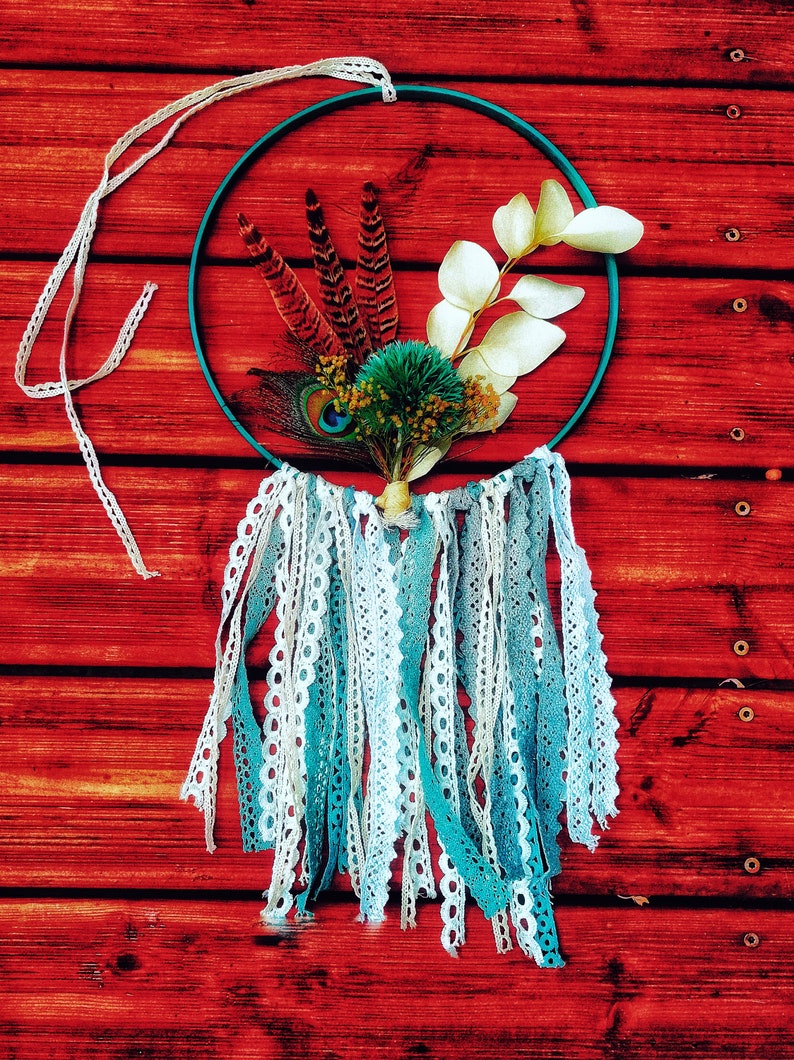 K&ouml;nnte beinhalten: Ein t&uuml;rkisfarbener Traumf&auml;nger mit wei&szlig;er und blauer Spitzenborte, der an einer roten Holzwand h&auml;ngt. Der Traumf&auml;nger ist mit Federn, getrockneten Bl&auml;ttern und einer kleinen gr&uuml;nen Bl&uuml;te verziert.