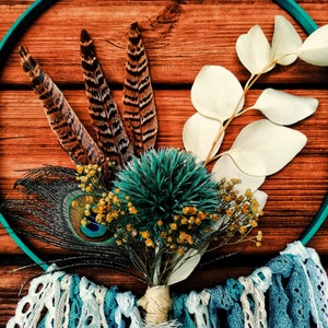 K&ouml;nnte beinhalten: Ein t&uuml;rkisfarbener Traumf&auml;nger mit wei&szlig;er Spitzenborte, verziert mit einem Strau&szlig; getrockneter Blumen, Pfauenfedern und braunen Fasanenfedern.