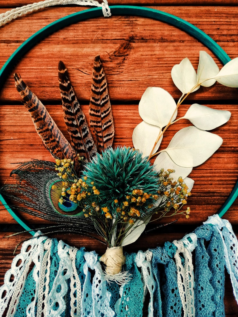 K&ouml;nnte beinhalten: Ein t&uuml;rkisfarbener Traumf&auml;nger mit wei&szlig;er Spitzenborte, verziert mit einem Strau&szlig; getrockneter Blumen, Pfauenfedern und braunen Fasanenfedern.