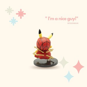 May include: A yellow Pikachu figurine wearing a red and blue Spiderman costume with gold web-like accents. The figurine is standing on a black base. The text "I'm a nice guy!" is written in pink above the figurine. The text "SPIDERMAN" is written in black below the figurine.