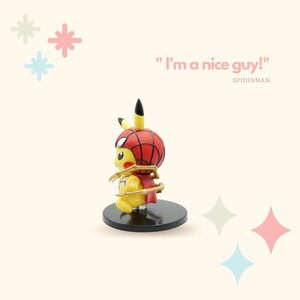 May include: A yellow Pikachu figurine wearing a red and blue Spiderman costume with a gold spider web design. The figurine is standing on a black base. The text "I'm a nice guy!" is written in pink above the figurine. The text "SPIDERMAN" is written in pink below the figurine.