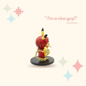 May include: A yellow Pikachu figurine wearing a red and blue Spiderman costume with gold web accents. The figurine is standing on a black base and the text "I'm a nice guy!" is written in pink above the figurine. The text "SPIDERMAN" is written in black below the figurine.