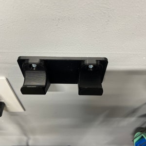 Peut inclure: Un support mural en plastique noir avec deux crochets pour accrocher des objets. Le support est fixé à un mur blanc avec des vis.