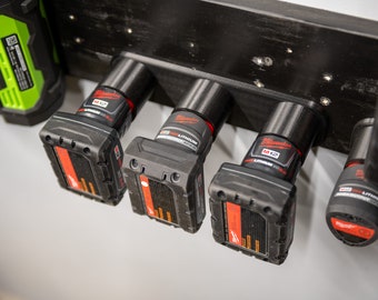 May include: Five black and red Milwaukee M12 Lithium-ion batteries are hanging on a black wall mount. The batteries are arranged in a row, with the battery on the left being the largest and the battery on the right being the smallest.