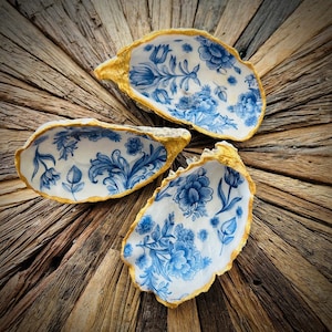 Peut inclure: Trois coquilles d'huîtres avec une bordure dorée et un motif floral bleu et blanc. Les coquilles sont disposées en cercle sur une surface en bois.