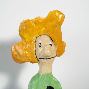 May include: A whimsical ceramic figurine with a yellow, cloud-like headpiece and a green body. The face is simple with black eyes and a smile. Two dark blue spots adorn the green garment. This unique art piece is a charming decorative item.
