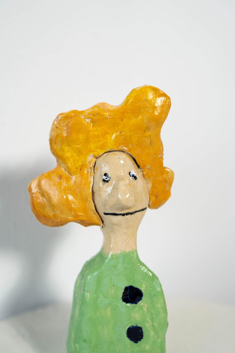 May include: A whimsical ceramic figurine with a yellow, cloud-like headpiece and a green body. The face is simple with black eyes and a smile. Two dark blue spots adorn the green garment. This unique art piece is a charming decorative item.