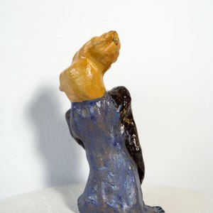 May include: A ceramic sculpture of a figure with a tan head and shoulders, a dark brown back, and a blue dress. The figure is seated, with the head tilted upwards. The sculpture is on a white surface.