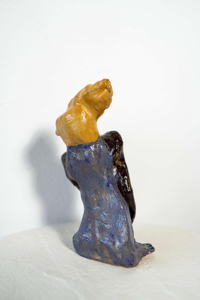 May include: A ceramic sculpture of a figure with a tan head and shoulders, a dark brown back, and a blue dress. The figure is seated, with the head tilted upwards. The sculpture is on a white surface.