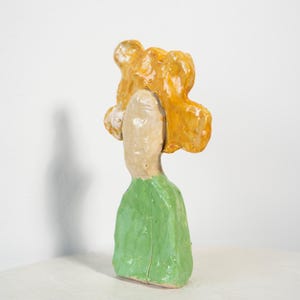 May include: A ceramic sculpture of a figure with a light beige face and a green garment. The figure has bright yellow hair. The sculpture is set against a white background.