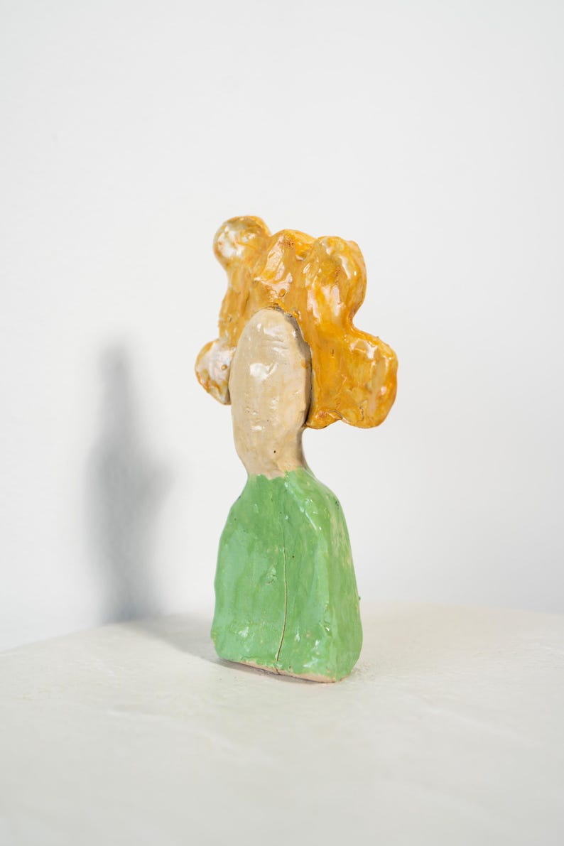 May include: A ceramic sculpture of a figure with a light beige face and a green garment. The figure has bright yellow hair. The sculpture is set against a white background.