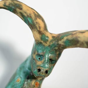 May include: A handcrafted sculpture of a creature with a face and horns. The sculpture has a weathered appearance with a teal and tan color palette. The face has a slightly sad expression. The sculpture is a unique piece of art.