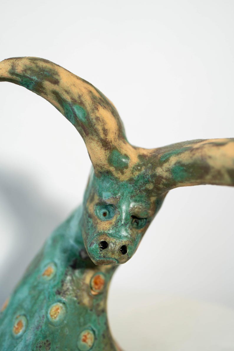 May include: A handcrafted sculpture of a creature with a face and horns. The sculpture has a weathered appearance with a teal and tan color palette. The face has a slightly sad expression. The sculpture is a unique piece of art.