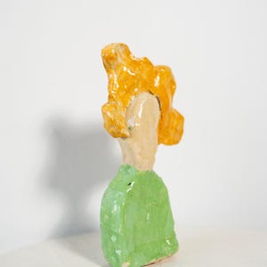 May include: A handmade ceramic sculpture of a figure with a green top and orange hair. The figure is painted with bright colors and has a unique, abstract design. The sculpture is set against a white background.