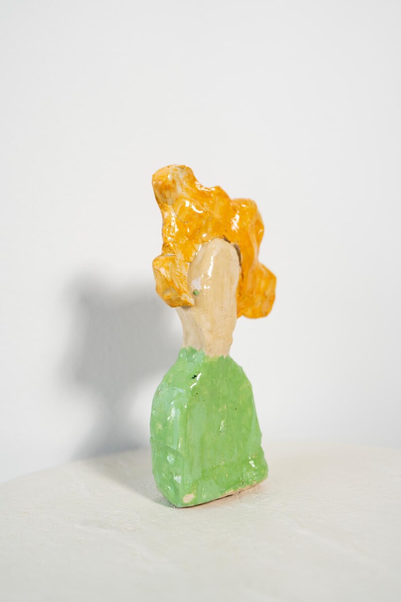 May include: A handmade ceramic sculpture of a figure with a green top and orange hair. The figure is painted with bright colors and has a unique, abstract design. The sculpture is set against a white background.
