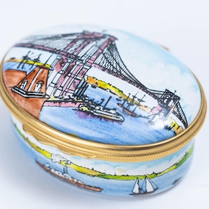 May include: Oval trinket box with a hand-painted illustration of a suspension bridge over water, likely the Brooklyn Bridge. The box has a gold-colored metal rim and a light blue background with boats.
