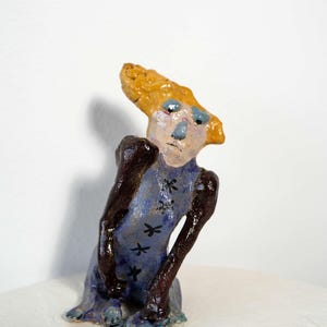 May include: A whimsical ceramic figurine with a stylized human form. The figure has a light blue dress adorned with black dragonfly designs, dark brown arms, and a yellow head. The face is painted with blue eyes and a light pink complexion.