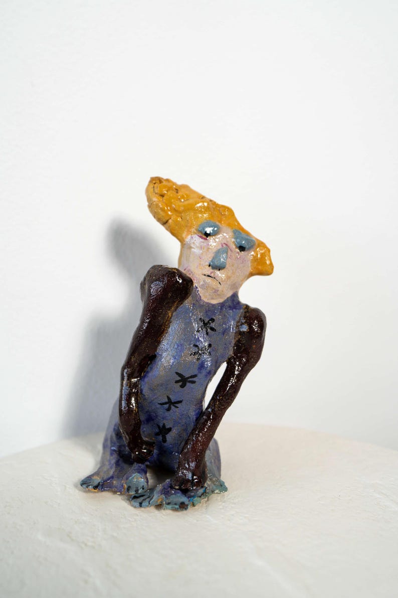 May include: A whimsical ceramic figurine with a stylized human form. The figure has a light blue dress adorned with black dragonfly designs, dark brown arms, and a yellow head. The face is painted with blue eyes and a light pink complexion.