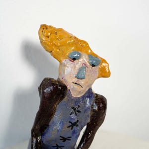 May include: A whimsical ceramic sculpture of a figure with a yellow head and blue face. The figure wears a blue garment with black cross-stitch details. The sculpture has a unique, handcrafted appearance.