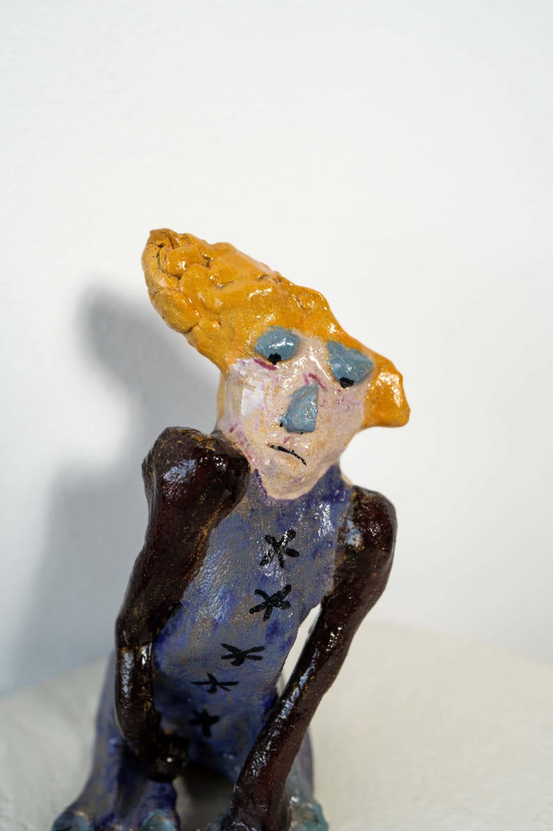 May include: A whimsical ceramic sculpture of a figure with a yellow head and blue face. The figure wears a blue garment with black cross-stitch details. The sculpture has a unique, handcrafted appearance.