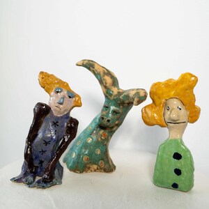 May include: Three whimsical ceramic figurines. One is a blue figure with a yellow head, another is a green creature with a curved neck, and the last is a figure with a yellow head and green dress. Each has a unique, expressive face.