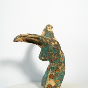 May include: A ceramic sculpture with a bird-like form. The sculpture features a tan beak and head, with a body in shades of teal and brown. The body is adorned with orange-brown circular spots. The artwork is displayed on a white surface.