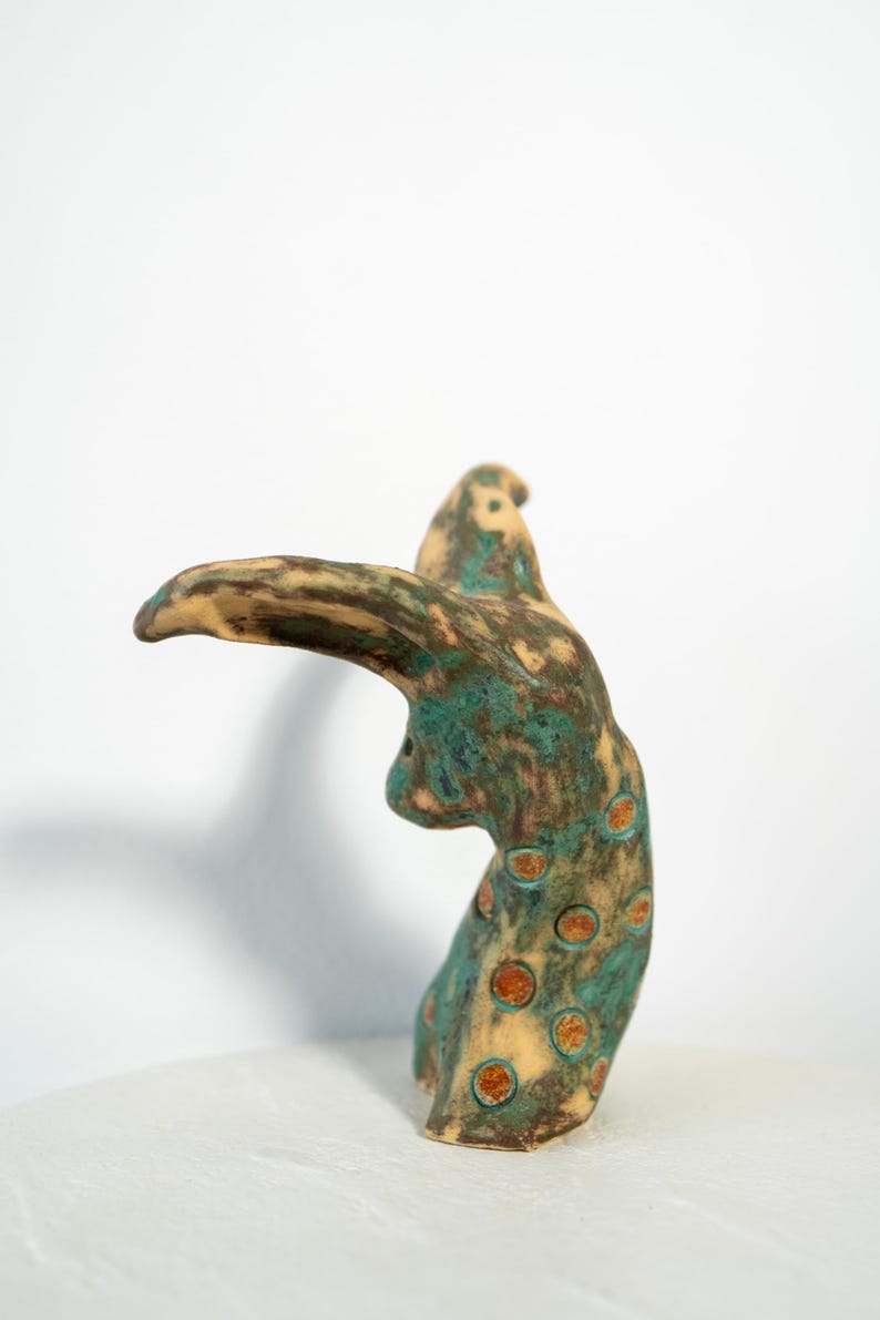 May include: A ceramic sculpture with a bird-like form. The sculpture features a tan beak and head, with a body in shades of teal and brown. The body is adorned with orange-brown circular spots. The artwork is displayed on a white surface.