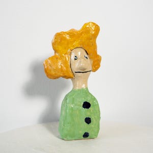May include: A whimsical ceramic figurine with a yellow, textured head and a green body adorned with three black dots. The face has simple, hand-painted features. The sculpture is set against a plain white background.