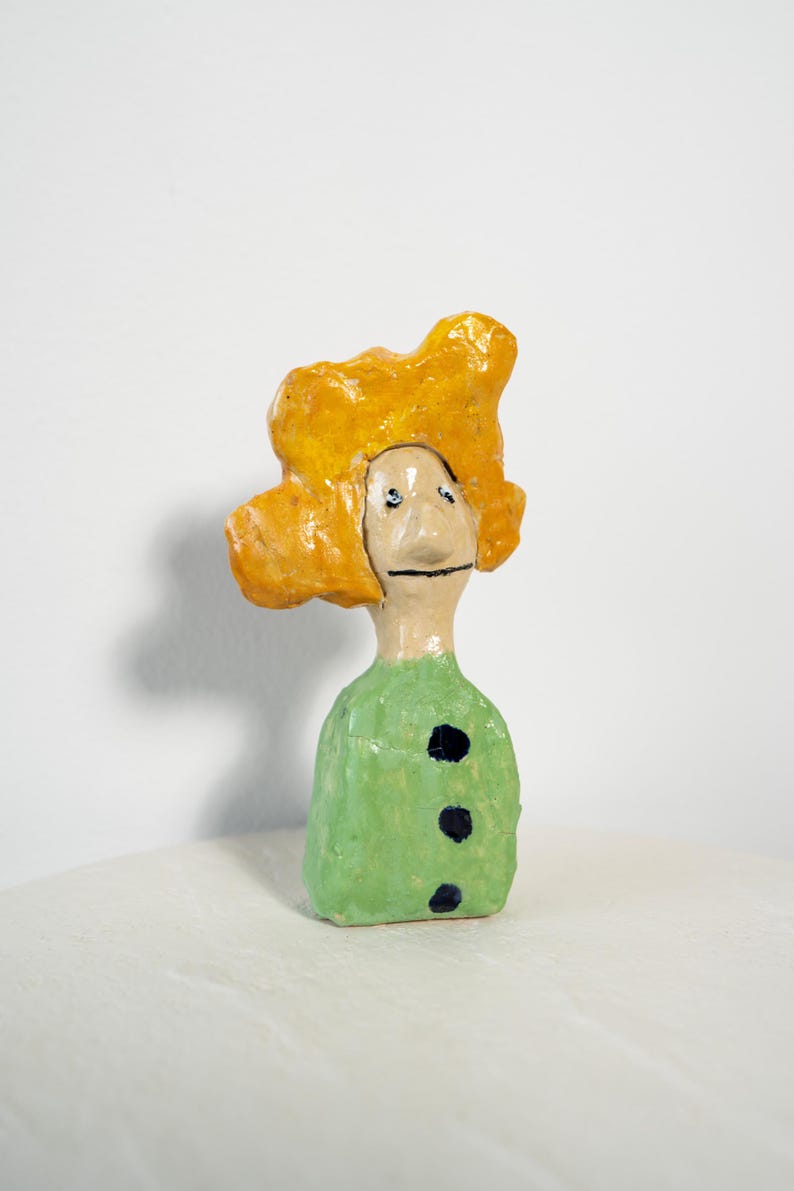 May include: A whimsical ceramic figurine with a yellow, textured head and a green body adorned with three black dots. The face has simple, hand-painted features. The sculpture is set against a plain white background.