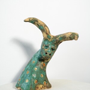 May include: A ceramic sculpture of a creature with a face and horns. The sculpture is primarily teal with orange and brown spots. The horns and the top of the head are tan. The sculpture is on a white surface.