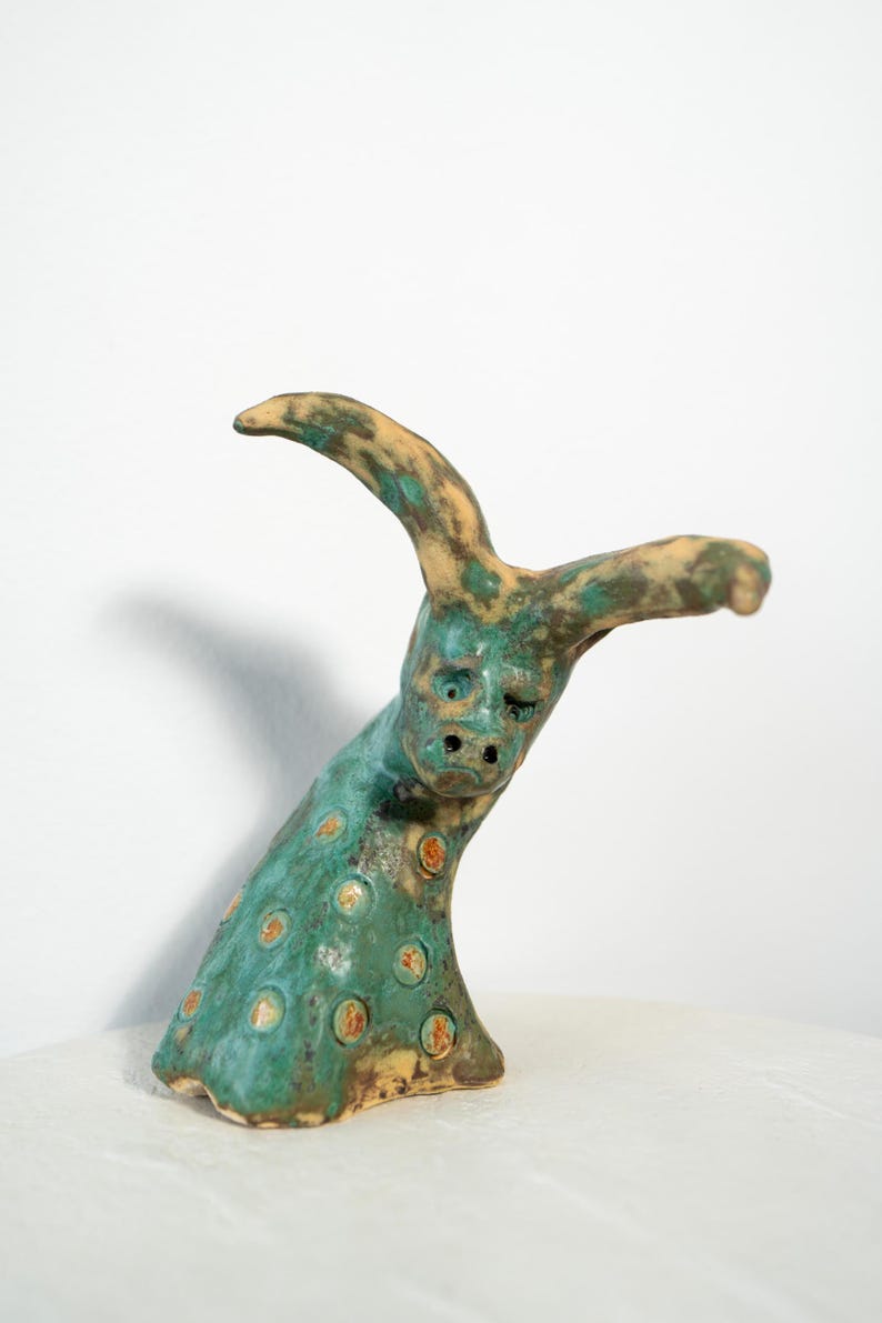 May include: A ceramic sculpture of a creature with a face and horns. The sculpture is primarily teal with orange and brown spots. The horns and the top of the head are tan. The sculpture is on a white surface.