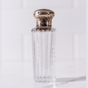 Pode incluir: Um frasco de perfume de vidro transparente com uma rolha prateada e dourada. A garrafa tem um design de nervuras verticais. A rolha é redonda com uma superfície texturizada. A garrafa está vazia e assenta numa superfície branca.
