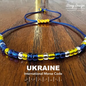 May include: A blue and yellow beaded necklace with a cord closure. The beads are arranged in a pattern representing the Ukrainian flag. The necklace is on a wooden surface. The text "UKRAINE" and "International Morse Code" is below the necklace. The Morse code for "UKRAINE" is shown below the text.