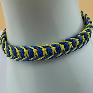 May include: A braided bracelet in a repeating pattern of blue and yellow. The bracelet is displayed on a white surface, showcasing the intricate weave and vibrant colors. The bracelet is made of a soft, flexible material.