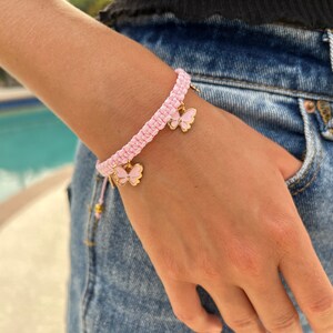 May include: A pink braided bracelet with two gold butterfly charms. The bracelet is worn on a person's wrist.