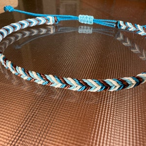 May include: A braided bracelet with blue, white, and brown cords. The bracelet is made with a simple knot pattern.