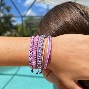 Puede incluir: Un conjunto de cuatro pulseras en tonos morados y rosados. Las pulseras están hechas de cuerda trenzada y tienen cuentas plateadas.