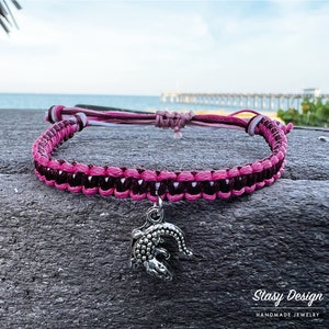 May include: A handmade macrame bracelet with a pink and dark brown woven pattern. A silver alligator charm hangs from the center. The bracelet is displayed on a gray surface with a blurred background of the ocean and a pier.