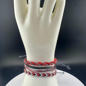 May include: A set of five bracelets in red, black, white, and gray. The bracelets are made of woven string and have a variety of patterns.
