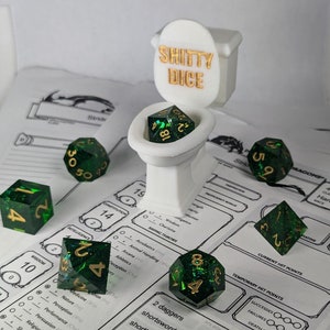 Könnte beinhalten: Ein Satz grüner und goldener Würfel mit glitzernder Oberfläche, die auf einem weißen, toilettenförmigen Halter liegen. Der Halter trägt die Aufschrift "Shitty Dice" in goldenen Buchstaben. Die Würfel befinden sich auf einem Tischspielbrett mit einem Charakterblatt.